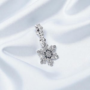 Puede incluir: Un charm de copo de nieve plateado con cristales transparentes. El charm cuelga de una fianza decorativa con pequeños adornos redondos. El diseño del copo de nieve tiene múltiples puntas, sobre fondo satinado blanco.