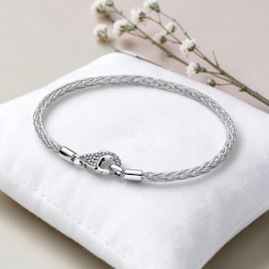 Op de afbeelding: Een zilveren armband met een geweven ontwerp, met een decoratieve sluiting met kleine, sprankelende accenten. De armband wordt gepresenteerd op een witte, zachte ondergrond, met delicate witte bloemen op de achtergrond.