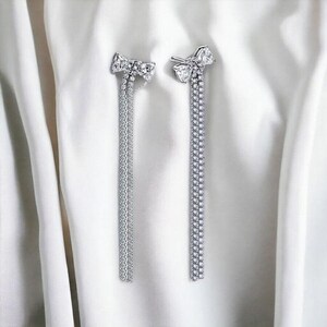 Puede incluir: Un par de pendientes plateados con forma de lazo y colgantes de pequeños cristales transparentes. Los pendientes se exhiben sobre un fondo de tela blanca drapeada, creando un contraste que resalta su brillo y diseño.