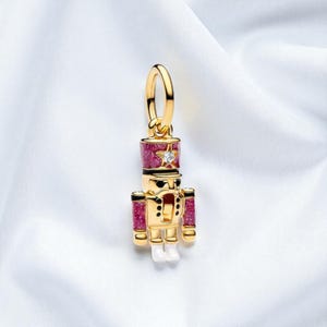 May include: A gold-colored charm in the shape of a nutcracker soldier. The charm features a red and glittery hat and accents, a star and a small diamond. The soldier has white boots and black details. The charm is suspended from a gold loop.