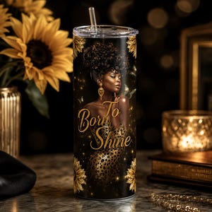 May include: A cylindrical tumbler with a clear lid and straw, featuring a black background with a graphic of a person with dark skin and curly hair, wearing gold earrings and a leopard print dress, with the gold text Born To Shine and sunflowers.