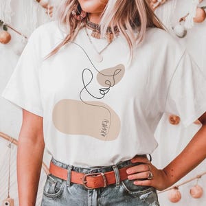 May include: White t-shirt with a brown abstract design and the word "love" written in cursive. The design includes a brown blob with the word "above" written inside.