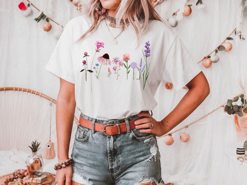 K&ouml;nnte beinhalten: Wei&szlig;es T-Shirt mit einem Blumendruck, der verschiedene Blumen in Rosa-, Lila- und Blaut&ouml;nen zeigt. Die Blumen sind in einer Reihe &uuml;ber die Vorderseite des Shirts angeordnet.