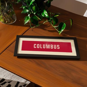 Puede incluir: Un marco de fotos rectangular negro que contiene una impresión horizontal con un fondo rojo sólido y la palabra COLUMBUS impresa en letras mayúsculas blancas. La impresión está centrada dentro del marco, que tiene un borde blanco.