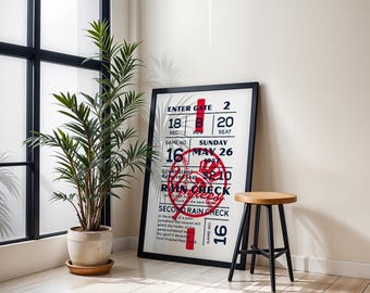 May include: A framed print with a black border featuring a baseball-themed design with red and blue accents. The print includes text such as "ENTER GATE", "RAIN CHECK", and "GAME NO 16". A wooden stool with black legs and a potted plant are also in the image.