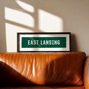 Puede incluir: Una pieza de arte mural rectangular enmarcada que presenta un panel horizontal verde oscuro con el texto blanco "EAST LANSING" impreso en una fuente sans-serif en negrita. La obra está encerrada en un marco negro fino sobre la pared.