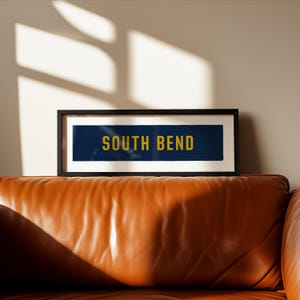 Puede incluir: Una impresión enmarcada rectangular que presenta un fondo azul oscuro con el texto SOUTH BEND impreso en letras mayúsculas amarillas en negrita. La obra está encerrada en un marco negro fino con un borde blanco, presentando un diseño limpio.