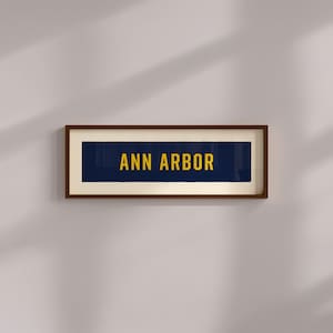 Puede incluir: Una pieza de arte mural rectangular que presenta una banda horizontal azul oscuro con el texto ANN ARBOR impreso en letras mayúsculas amarillas en negrita. La obra está encerrada en un marco de madera marrón oscuro con un borde crema.