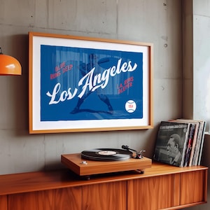 Puede incluir: Una impresión enmarcada con un fondo azul y las palabras "Los Angeles" en escritura blanca. Se ve una silueta de jugador de béisbol. La impresión está en un marco de madera, exhibida sobre un tocadiscos de madera y discos de vinilo.