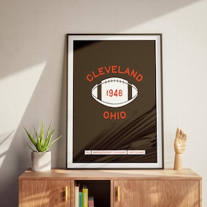 Puede incluir: Un póster en blanco y negro con un fondo marrón que presenta un balón de fútbol blanco con un "1946" rojo en su interior. El texto "CLEVELAND OHIO" está en rojo sobre el balón y el texto "NFL AMERICAN FOOTBALL CONFERENCE NORTH DIVISION" está en blanco debajo del balón.