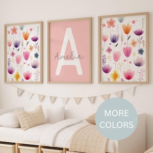 May include: Three framed floral art prints with watercolor flowers in pink, purple, orange, and yellow. The center print features a large white letter "A" and the name "Amelia" on a pink background. The frames are light brown.