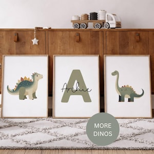 Könnte beinhalten: Drei gerahmte Kunstdrucke mit Dinosaurier-Illustrationen. Einer zeigt einen grünen und blauen Stegosaurus, ein anderer zeigt den Buchstaben "A" mit dem Namen "Archie", und der dritte zeigt einen Brontosaurus. Die Drucke werden auf einem Holzschrank ausgestellt.