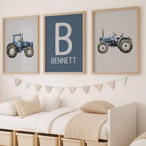 Könnte beinhalten: Drei gerahmte Drucke mit Bauernhof-Motiv. Zwei Drucke zeigen blaue Aquarelltraktoren auf hellen Hintergründen. Der mittlere Druck hat ein großes weißes "B" und den Namen "BENNETT" auf blauem Hintergrund. Die Rahmen sind aus hellem Holz.