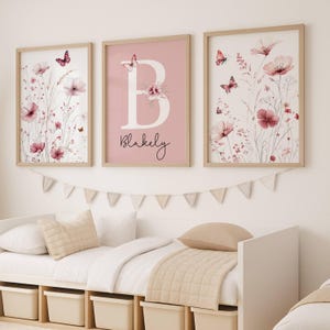 May include: Three framed floral art prints with a light wood frame. The prints feature pink flowers, butterflies, and the name "Blakely" with the letter "B". The prints are displayed above a white bed with beige bedding.