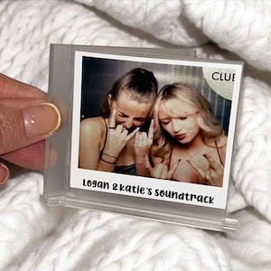 May include: A white photo frame with a Polaroid-style photo of two women making the rock and roll hand gesture. The photo is titled "Logan & Katie's soundtrack" and has the word "CLUE" written in the top right corner.