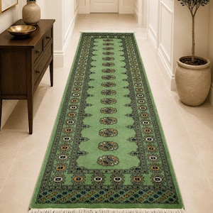 Puede incluir: Una alfombra pasillera larga y rectangular en un tono verde salvia suave, con una columna central de medallones geométricos repetidos en azul, oro y negro. El borde muestra patrones intrincados, rematado con flecos blancos en ambos extremos.