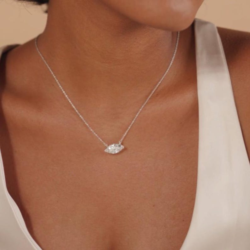 May include: A delicate silver necklace featuring a marquise-cut diamond pendant. The pendant is the focal point, suspended from a fine chain. The necklace is worn against a skin tone, highlighting the jewelry's sparkle and elegance.