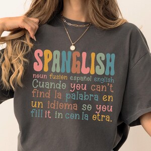 May include: Dark gray t-shirt with the word "SPANGLISH" in colorful, retro-style lettering. The shirt features text in English and Spanish, discussing the fusion of the two languages. The shirt is a relaxed fit.