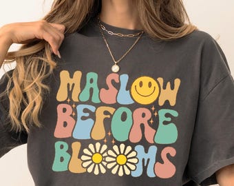 May include: Dark gray t-shirt with the text "MASLOW BEFORE BLOOMS" in colorful retro font. The design includes a yellow smiley face and daisy flowers. The shirt is paired with blue jeans and black sneakers.