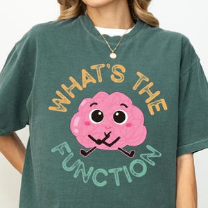 May include: A teal-colored t-shirt with the phrase "WHAT'S THE FUNCTION" and a cartoon brain graphic. The brain is pink with black eyes and a smiling expression. The shirt is a casual, unisex style.