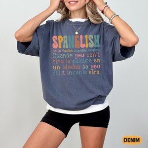 May include: A denim-colored t-shirt with the word "SPANGLISH" in colorful letters. Below the word is text in English and Spanish. The shirt has short sleeves and a relaxed fit.