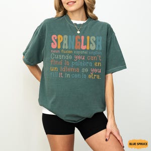 May include: A teal-colored t-shirt with the word "SPANGLISH" in colorful letters. Below, text in English and Spanish explains the concept of Spanglish. The shirt is a relaxed fit, suitable for casual wear.