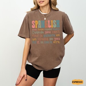 May include: A brown t-shirt with the word "SPANELISH" in colorful letters. The shirt has text below that reads "noun fusion espa&ntilde;ol english" and other phrases in English and Spanish. The shirt is a relaxed fit.