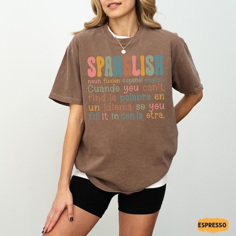 May include: A brown t-shirt with the word "SPANELISH" in colorful letters. The shirt has text below that reads "noun fusion espa&ntilde;ol english" and other phrases in English and Spanish. The shirt is a relaxed fit.