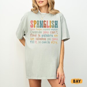 May include: A light green t-shirt with the word "SPANGLISH" in colorful letters. Below the word are additional lines of text in English and Spanish. The shirt has short sleeves and a relaxed fit.
