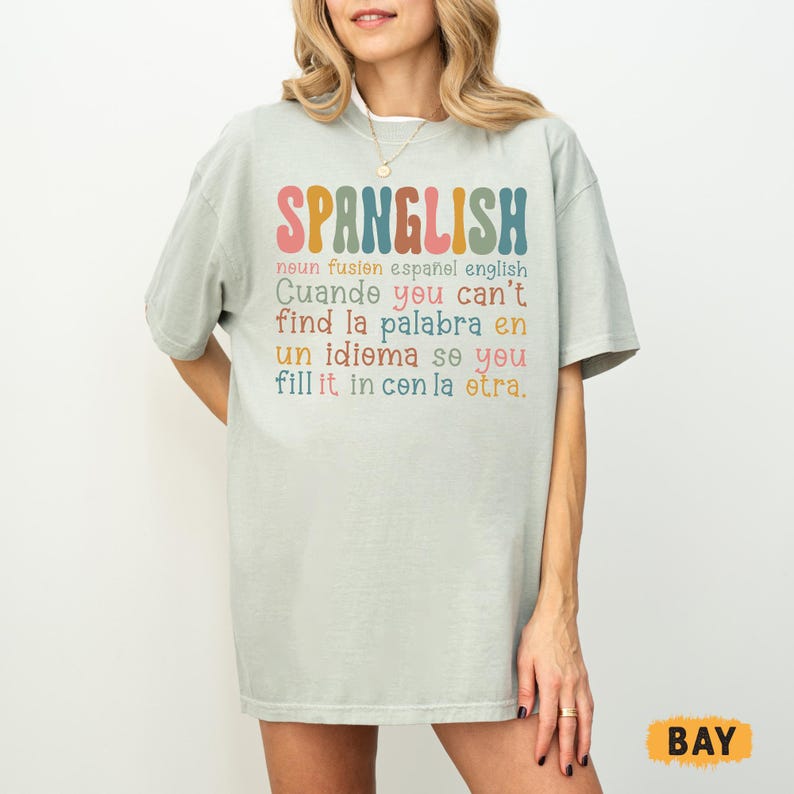 May include: A light green t-shirt with the word "SPANGLISH" in colorful letters. Below the word are additional lines of text in English and Spanish. The shirt has short sleeves and a relaxed fit.