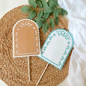 May include: Two decorative paper toppers with scalloped edges and floral cutouts. One is brown with white accents, the other is white with blue accents. Both have a blank rectangular space and are attached to white sticks.