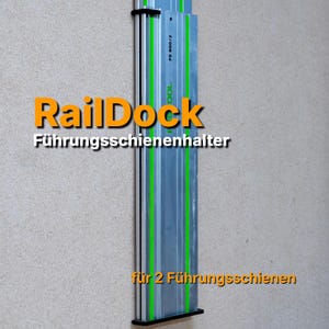 Op de afbeelding: Een zilver- en groenkleurige RailDock Führungs schienenhalter, ontworpen om twee geleiderails te bevatten. De tekst "RailDock Führungs schienenhalter" staat in oranje en wit. De geleiderailhouder is op een getextureerde muur gemonteerd.