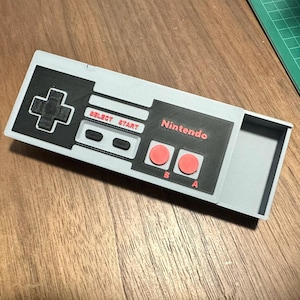 Peut inclure: Une manette de jeu Nintendo grise et noire avec des boutons rouges. La manette a un compartiment à l'arrière qui peut être ouvert.
