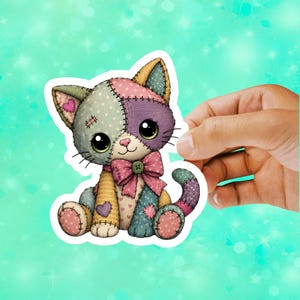 May include: A die-cut sticker featuring a patchwork cat illustration with large black eyes, a pink bow with a green button, and various patterned fabric textures in shades of sage green, purple, pink, and yellow with visible stitched seams.