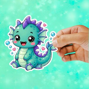 May include: A die-cut sticker featuring a teal dragon with purple dorsal fins, large black eyes, and a light green belly. The dragon is surrounded by small pink hearts and blue bubbles, rendered in a vibrant, colorful, and detailed illustration style.