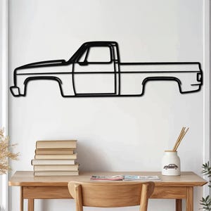 May include: Black metal silhouette of a pickup truck, mounted on a white wall. The truck is outlined in black and has a simple design. The truck is displayed against a white wall with a wooden table and chair in the foreground.