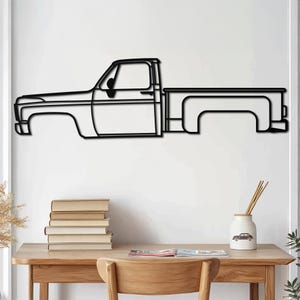 Puede incluir: Arte de pared de metal negro con la silueta de una camioneta vintage. La camioneta está delineada en negro y tiene un diseño simple. El arte está montado en una pared blanca sobre una mesa de madera con libros y un jarrón.