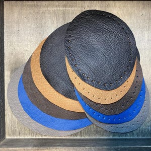 Puede incluir: Un conjunto de siete círculos de cuero en varios tonos de gris, azul, marrón y negro. Los círculos están apilados uno encima del otro y tienen un borde cosido.