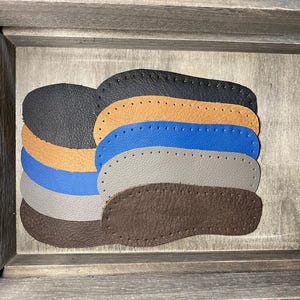 Könnte beinhalten: Ein Stapel aus fünf ovalen Ledereinlegesohlen mit vorgestanzten Löchern entlang der Ränder. Die Farben umfassen Schwarz, Hellbraun, Blau, Grau und Dunkelbraun. Jede Einlegesohle weist eine strukturierte Oberfläche sowie eine abgerundete Form auf.