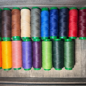 Könnte beinhalten: Eine Sammlung von einundzwanzig Spulen mit gewachstem Garn, die in zwei Reihen angeordnet sind. Die Spulen weisen eine Vielzahl von Volltonfarben auf, darunter Schwarz, Braun, Beige, Grau, Blau, Rot, Kastanienbraun, Orange, Gelb, Rosa, Lila, Limettengrün und Dunkelgrün.