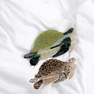 May include: Two decorative hair clips shaped like sea turtles. One clip is green and white with gold accents, the other is brown and tan with a tortoiseshell pattern. Both clips are on a white background.
