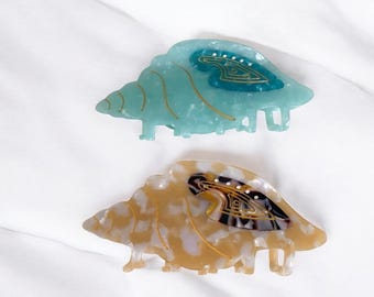 May include: Two tortoise shell hair clips in the shape of a sea shell. The top clip is teal with gold accents and the bottom clip is brown with gold accents.