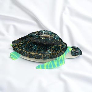 May include: A turtle-shaped clutch purse with a hinged top. The body is a mosaic of dark green and gold, with light green and blue accents on the flippers. The purse is set against a white background.