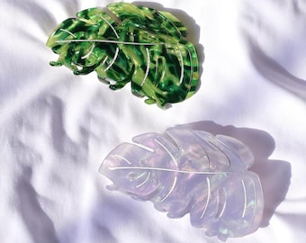 May include: Two plastic hair clips shaped like monstera leaves. One clip is green with white veins and the other is iridescent purple with white veins.