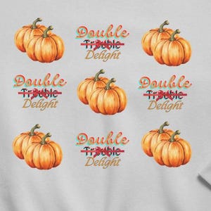 Può includere: Una felpa grigio chiaro con zucche ad acquerello e la scritta "Double Trouble Delight" in un font a tema autunnale. Il design include coppie di zucche arancioni. La felpa è circondata da foglie autunnali e decorazioni.
