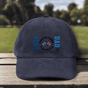 May include: Navy blue corduroy baseball cap with embroidered text. The text reads "TWIN DAD" with "Twice the Cuddles, Twice the Fun" and the year "2025" in a circular design. The cap has a curved brim.