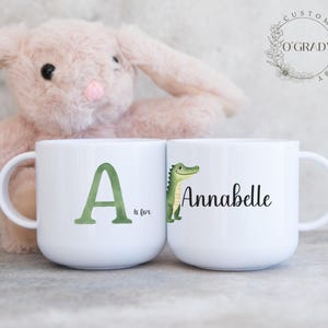 May include: Two white ceramic mugs. One mug features a large green letter "A" with "is for" below it. The other mug has the name "Annabelle" and a green cartoon alligator. The mugs are personalized gifts.