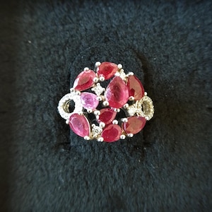 Op de afbeelding: Een zilveren ring met een cluster van rode en roze edelstenen, gezet in een zilveren band. De ring is omgeven door kleinere witte edelstenen.