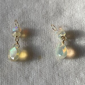 May include: A pair of gold-toned earrings featuring iridescent opal gemstones. Each earring has a small cluster of opals at the top, connected to a larger, teardrop-shaped opal below. The opals display a play of color.