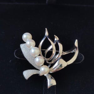 May include: A silver-toned brooch featuring a floral design with four white pearls. The brooch has a textured, ribbon-like element and a stylized, openwork design. The pearls are round and evenly spaced.